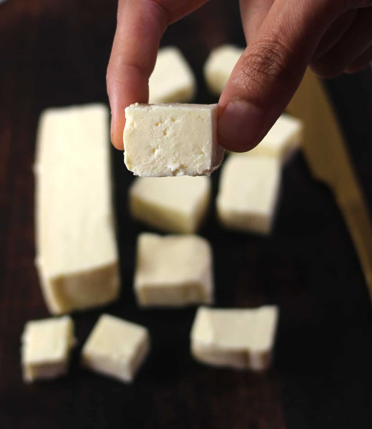 Holding a cube of fresh and soft homemade paneer - detailed steps on how to make paneer. 