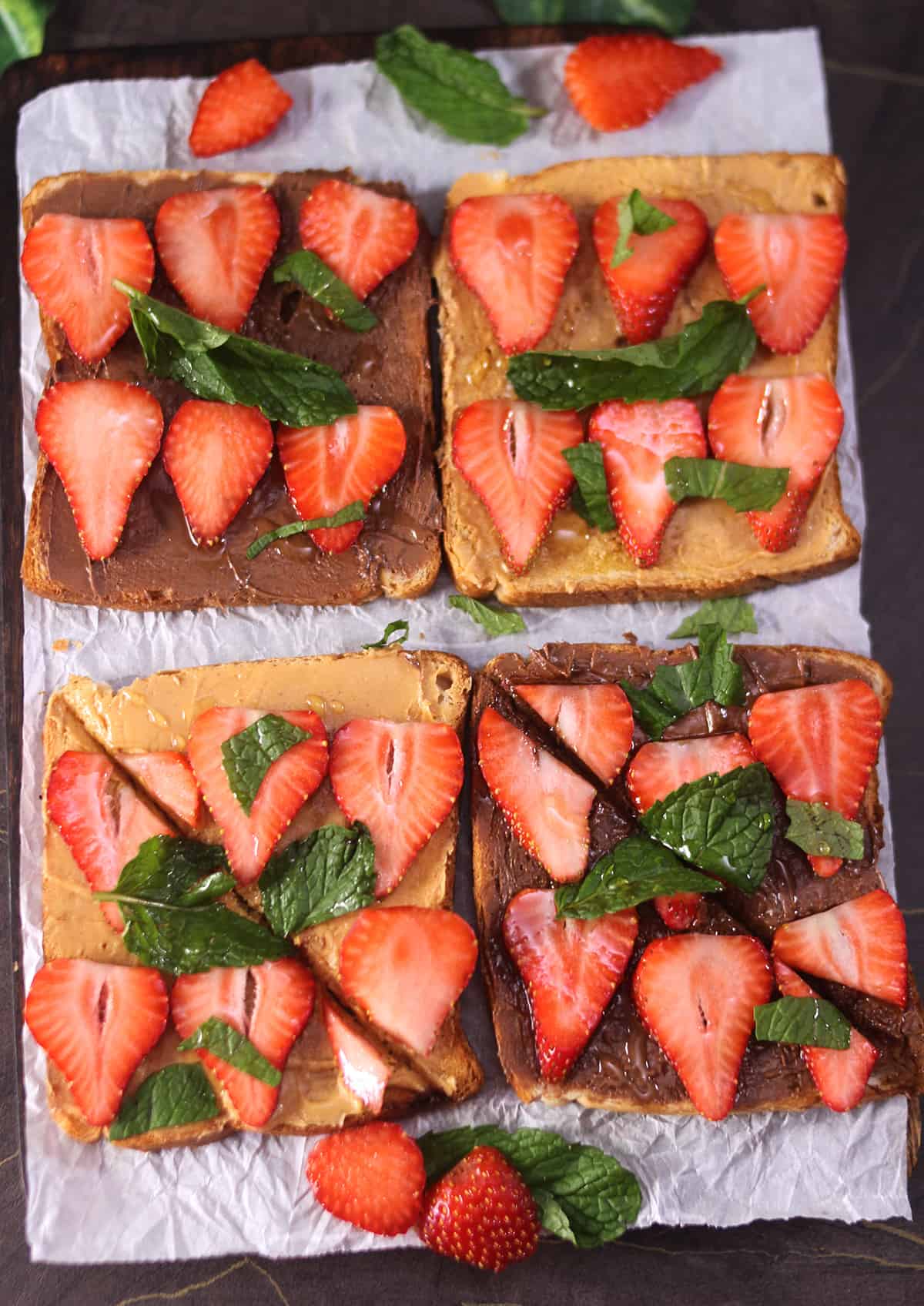 Strawberry toast with peanut butter and nutella placed on parchment paper, garnished with mint leaves. 