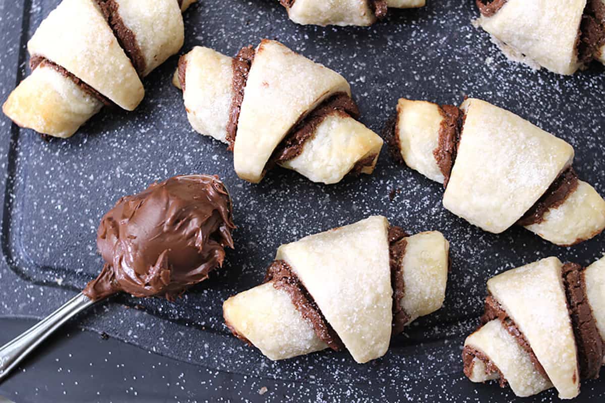 Top view of easy baked Nutella roll-ups with tablespoon of Nutella chocolate spread on a black plate. 