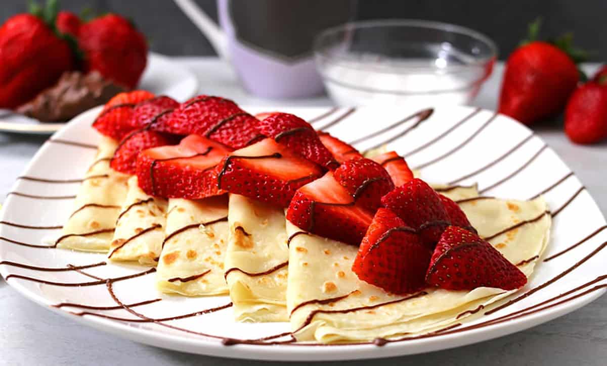 Best strawberry Nutella crepes on white serving plate with fresh berries and coffee in the background. 