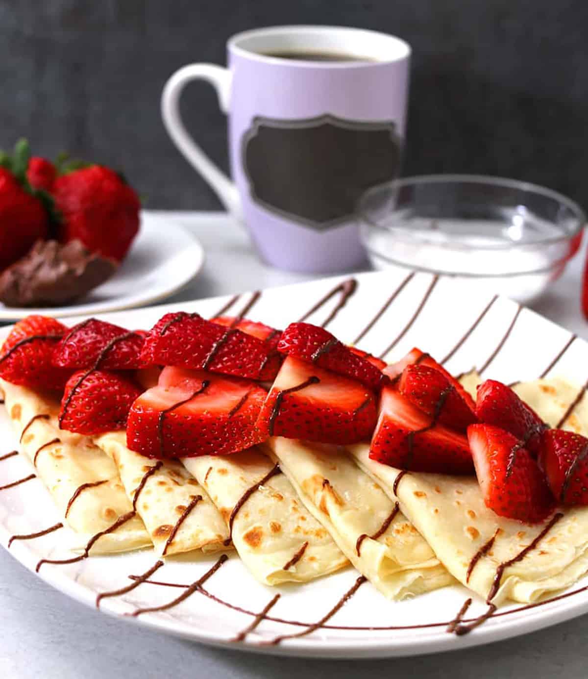 French Nutella Strawberry crepes on serving plate with coffee mug in background. 