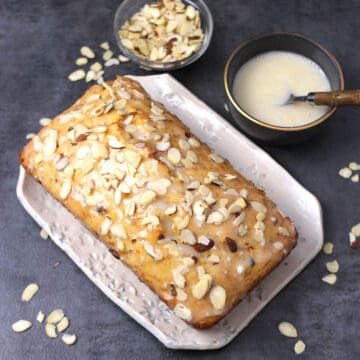 A loaf of carrot halwa/pudding cake served on a white rectangular plate, drizzle with a sour cream glaze and topped with sliced toasted almonds.
