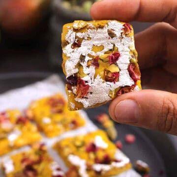 Holding a piece of mango burfi prepared with coconut and milk powder. - Indian barfi sweet.
