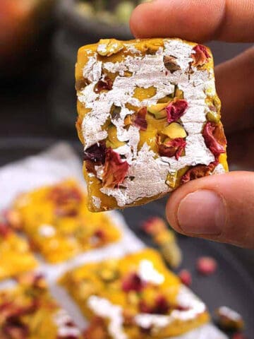Holding a piece of mango burfi prepared with coconut and milk powder. - Indian barfi sweet.