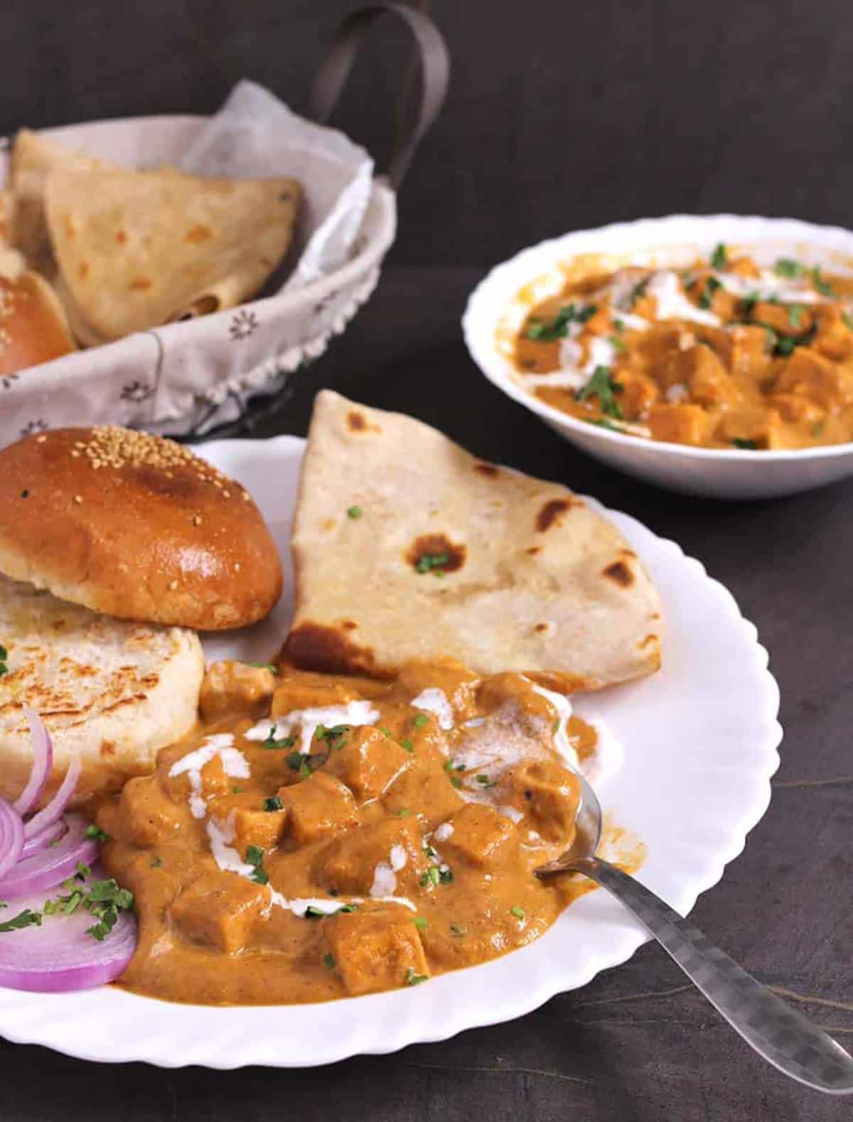 Butter paneer, toasted buns, chapati, and onion rings served in a plate. Butter paneer is garnished with cilantro and few spashes of cream. Background consists of a bread basket and serving bowl containing butter paneer.