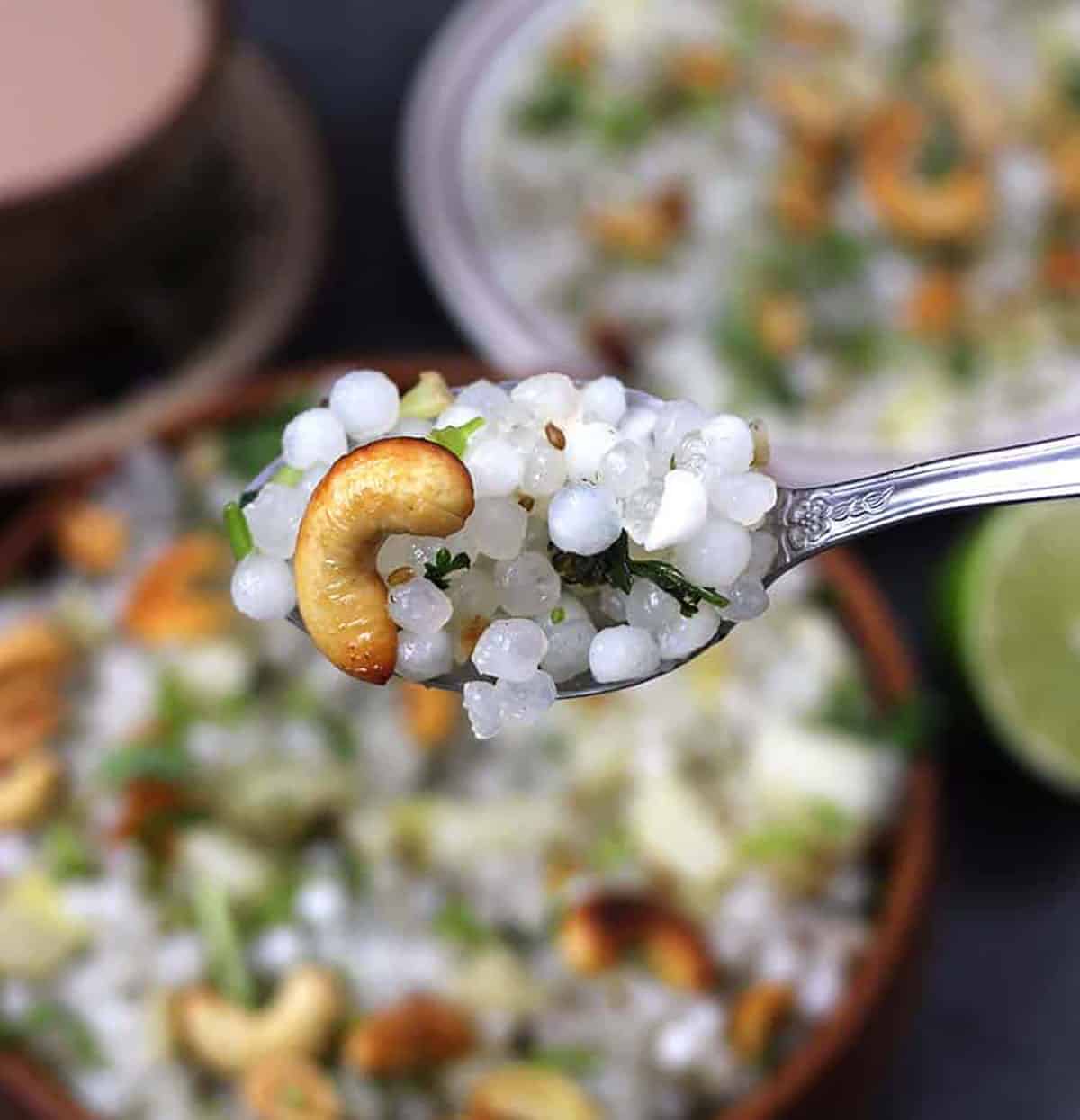 Top view of sabudana khichdi in a spoon, with bowl full of sabudana khichdi, which is out of focus,in the background.