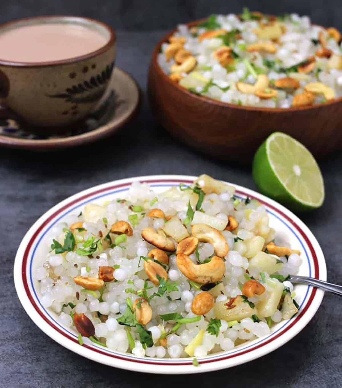 Plate of sabudana khichdi garnished with roasted cashews and peanuts, with a cut of tea, and half of a lime.