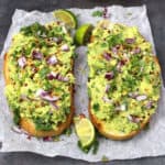 Two large avocado toasts topped with onion slices, red pepper flakes, and chopped cilantro. Lemon wedges are provided with the toasts.