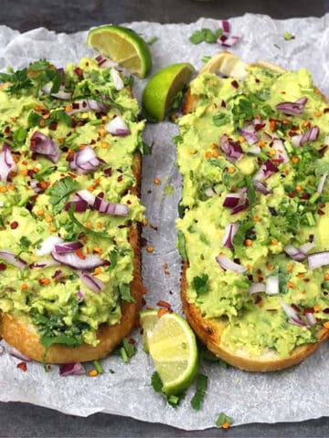 Two large avocado toasts topped with onion slices, red pepper flakes, and chopped cilantro. Lemon wedges are provided with the toasts.