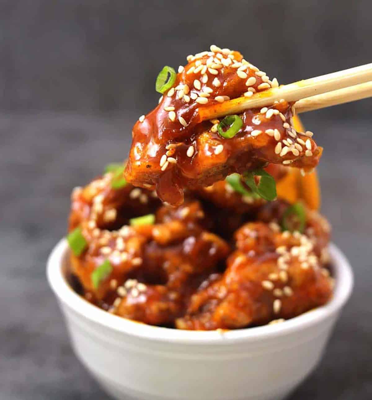 Orange chicken garnished with scallions and toasted sesame seeds picked using a pair of chopsticks. A white bowl containing orange chicken in the background.