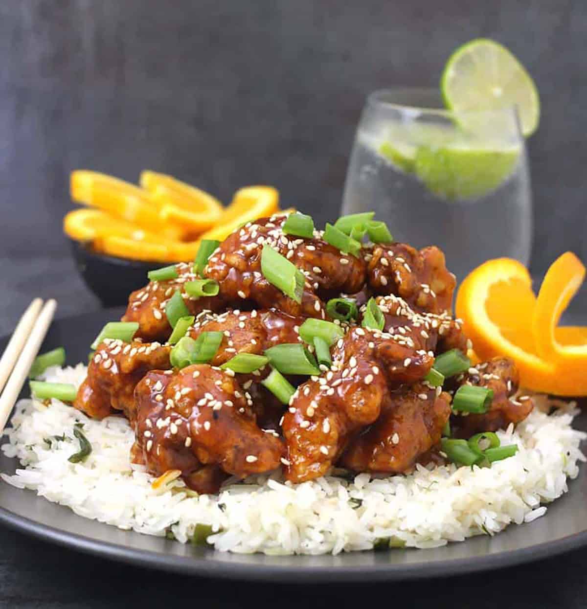 Orange chicken garnished with scallions and toasted sesame seeds served over a bed of steamed rice. In the background there is a glass of sparking water with lemon and orange wedges.