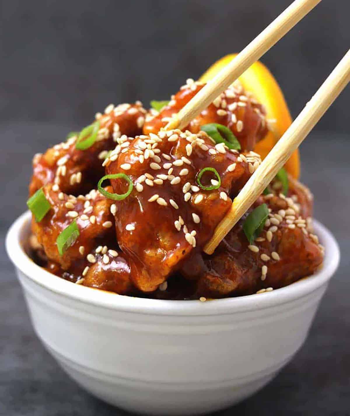 A white bowl containing orange chicken garnished with scallions and toasted sesame seeds. A piece of chicken is about to be picked using a pair of chopsticks.