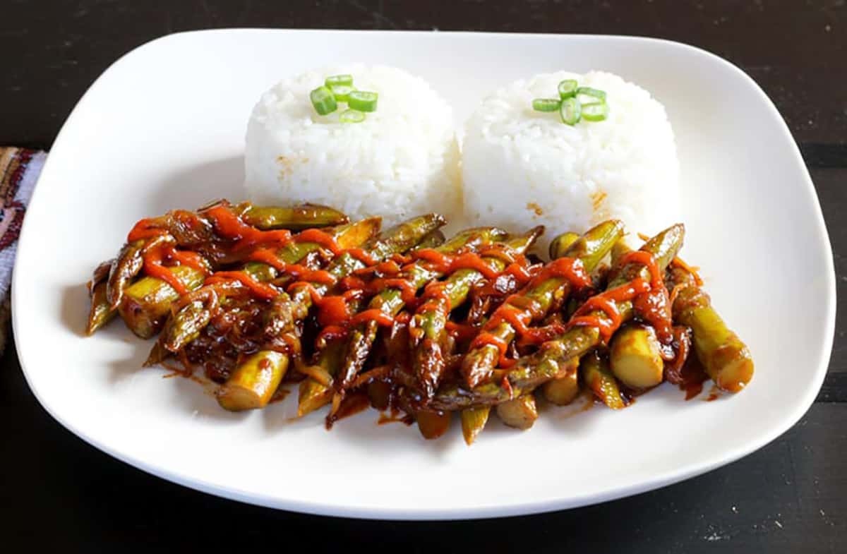 Stir Fried Asparagus with a drizzle of schezwan sauce served in a white plate with rice.
