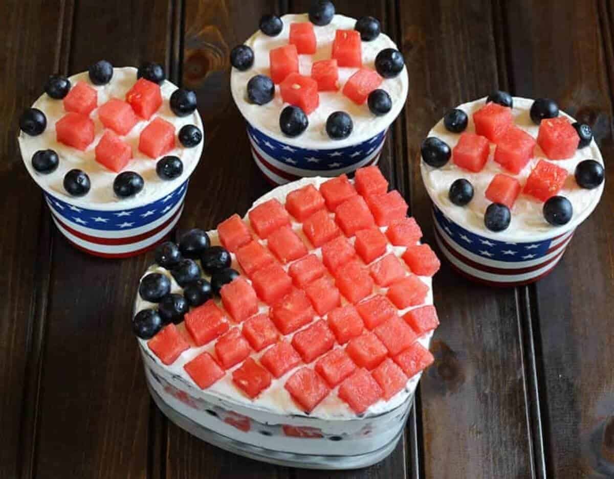 Simple and Best fruit and cream dessert served in a heart shaped glass bowl and paper ice cream cups. Watermelon cubes and blueberries are arranged as stripes and stars of the American flag.