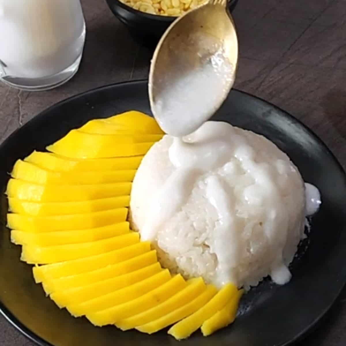 Pour salted coconut sauce over a small portion of sticky rice placed in a black plate containing sliced mangoes.