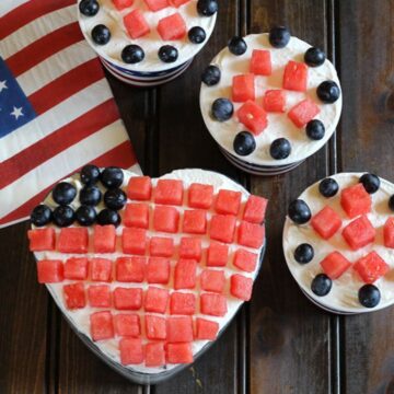 Easy fruit and cream dessert served in a heart shaped glass bowl and paper ice cream cups. Watermelon cubes and blueberries arranged stripes and stars of the American flag.