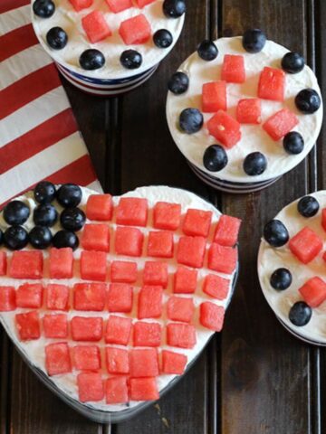 Easy fruit and cream dessert served in a heart shaped glass bowl and paper ice cream cups. Watermelon cubes and blueberries arranged stripes and stars of the American flag.