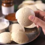 Holding healthy quinoa idli or millet idli with more idlis, sambar and chutney in background.