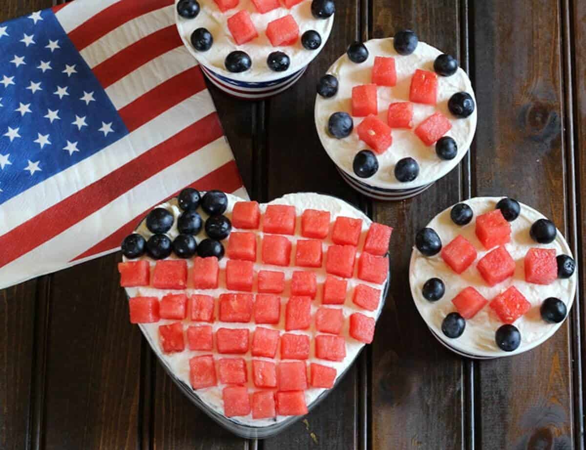 Easy and Best fruit with cream dessert served in a heart shaped glass bowl and paper sundae cups. Watermelon cubes and blueberries are arranged as stripes and stars on the top to resemble the American flag.