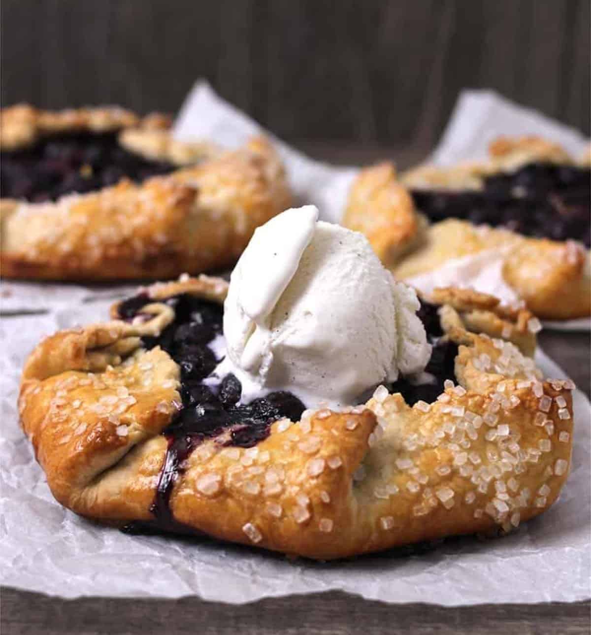 Side view of must-try blueberry galettes or crostatas on white parchment paper topped with a scoop of ice cream.
