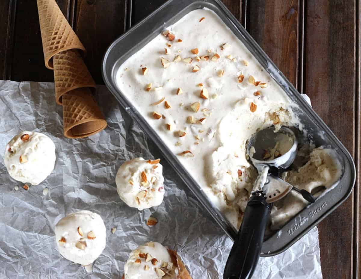 Scoops of roasted almond ice cream placed over parchment paper. A loaf pan containing frozen ice cream and  a scoop is in the background.
